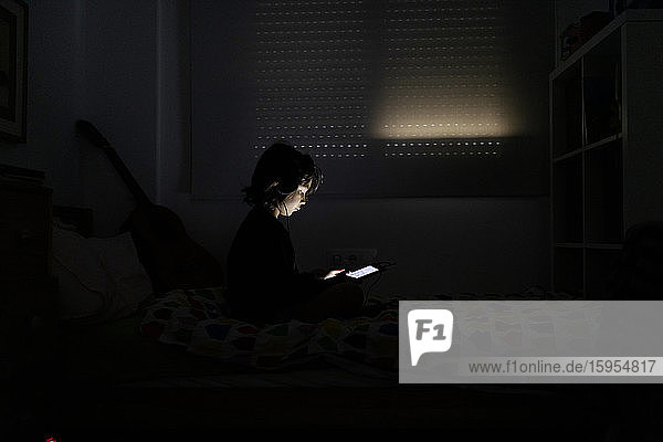 Junge benutzt Kopfhörer und digitales Tablet in seinem dunklen Zimmer zu Hause