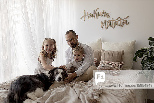 Porträt eines glücklichen Vaters  der mit seinen beiden Kindern auf dem Bett sitzt und den Hund streichelt