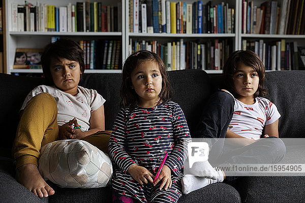 Süße Geschwister sehen fern  während sie auf dem Sofa vor dem Bücherregal im heimischen Wohnzimmer sitzen