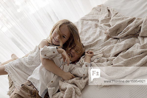 Porträt eines kleinen Mädchens  das mit seinem lachenden Bruder im Bett kuschelt