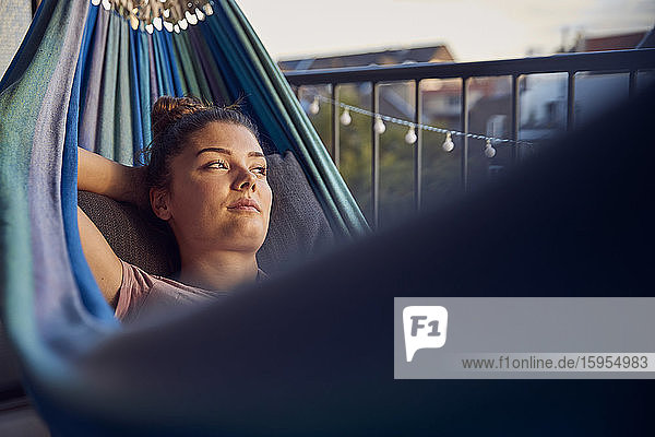 Porträt einer nachdenklichen jungen Frau  die auf einer Hängematte auf dem Balkon liegt und in die Ferne schaut