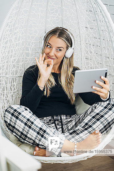 Portrait of woman sitting on a swinging chair at home using mini tablet and headphones for video chat