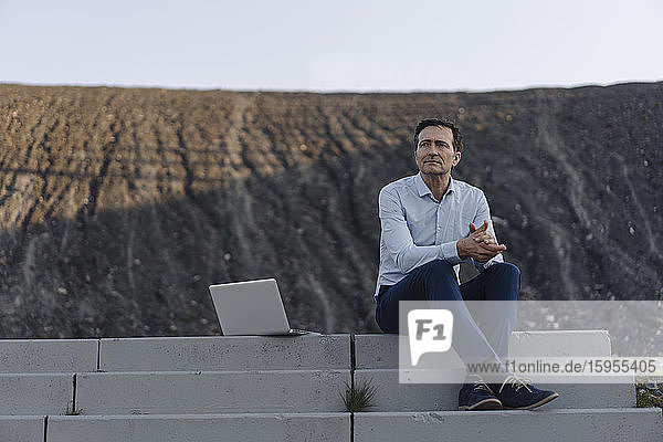 Ein reifer Geschäftsmann sitzt auf einer Treppe auf einer stillgelegten Minenhalde neben einem Laptop