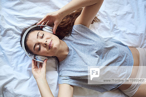 Porträt einer jungen Frau mit geschlossenen Augen auf dem Bett liegend und mit Kopfhörern Musik hörend