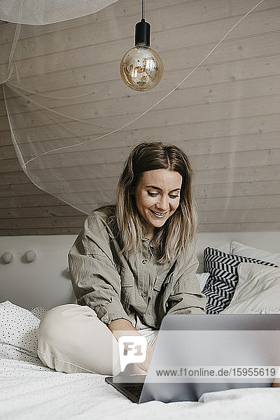 Lächelnde Frau benutzt Laptop  während sie zu Hause auf dem Bett sitzt