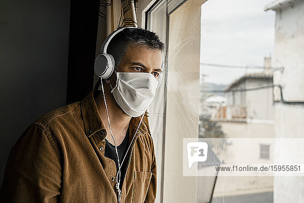 Mann mit Schutzmaske und weißen Kopfhörern  der aus dem Fenster schaut