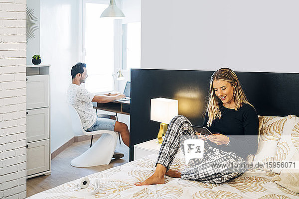 Lächelnde Frau sitzt im Bett und benutzt ein digitales Tablet  während ihr Freund im Hintergrund am Laptop arbeitet