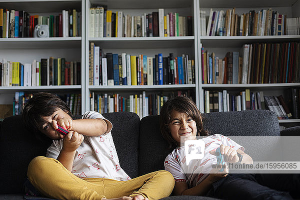 Geschwister spielen Videospiel  während sie zu Hause auf dem Sofa gegen ein Bücherregal im Wohnzimmer sitzen