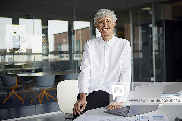 Porträt einer lächelnden älteren Geschäftsfrau  die in ihrem Büro auf dem Schreibtisch sitzt