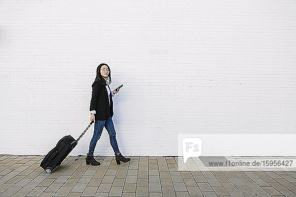 Young businesswoman with mobile phone and trolley bag walking in front of white wall