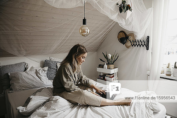 Junge Frau benutzt Laptop  während sie zu Hause auf dem Bett sitzt
