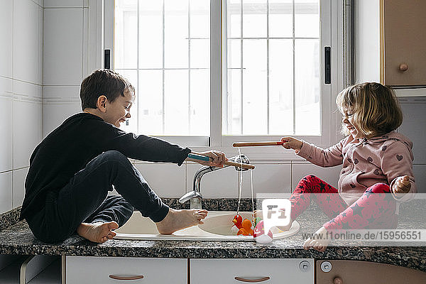 Boy and his little sister fishing rubber ducks in kitchen sink