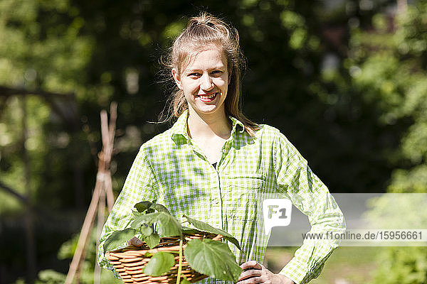 Porträt einer lächelnden schönen jungen Frau  die einen Weidenkorb hält  während sie an einem sonnigen Tag im Garten steht