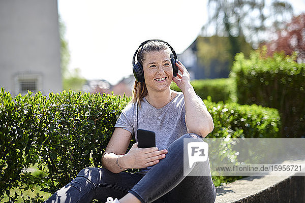 Lächelnde Frau schaut weg  während sie über Kopfhörer am Smartphone bei Pflanzen Musik hört