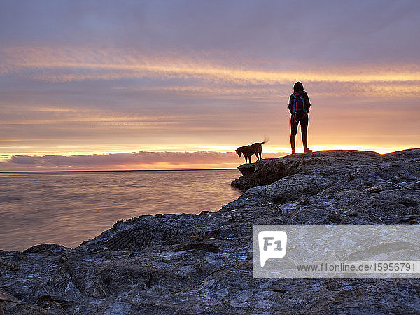 Rückansicht einer Frau  die mit einem Hund auf einer Felsformation auf dem Meer bei Sonnenuntergang steht  Puerto Piramides  Argentinien