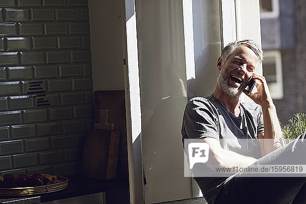 Laughing mature man sitting at the window at home talking on the phone