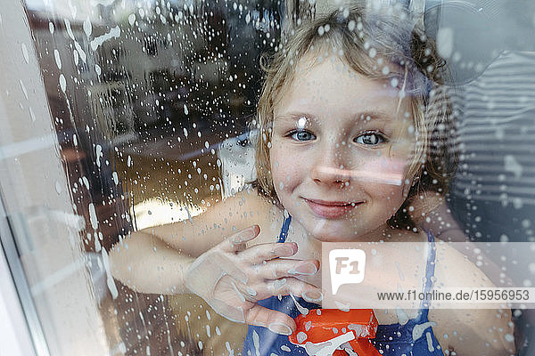 Porträt eines lächelnden blonden Mädchens mit Sprühflasche  das durch ein nasses Fenster schaut