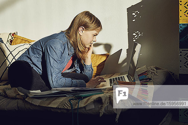 Girl sitting on bed at home doing homework and using laptop