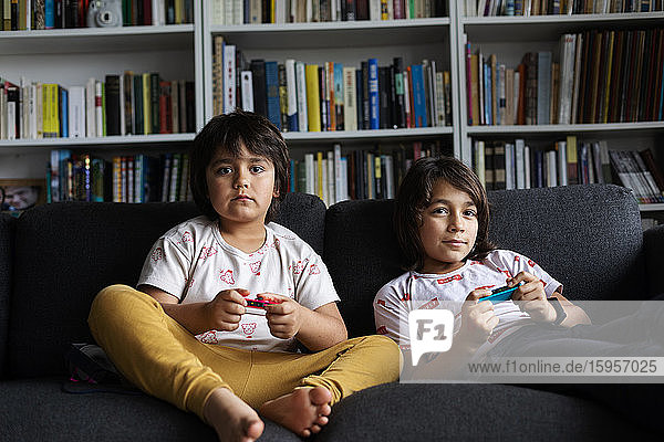 Brothers playing video game while sitting on sofa against bookshelf in living room at home