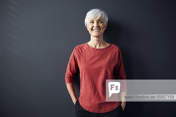 Porträt einer lächelnden älteren Frau in rotem Hemd vor grauer Wand stehend