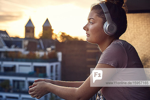 Junge Frau  die bei Sonnenuntergang auf dem Balkon mit Kopfhörern Musik hört