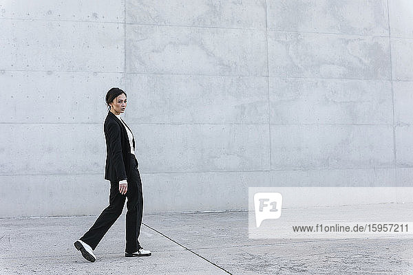 Junge Frau in schwarzem Anzug geht vor einer Betonmauer