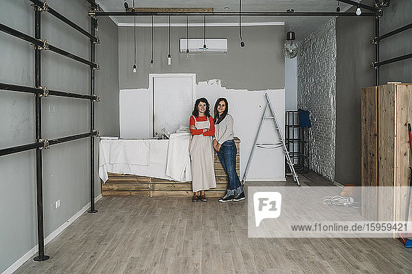 Zwei mittelgroße erwachsene Frauen  die sich in ihrem neuen Laden an einen Tisch lehnen  Porträt in voller Länge