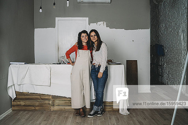 Zwei mittelgroße erwachsene Frauen lehnen gegen einen Tisch  während sie in ihrem neuen Laden malen  Ganzkörperporträt