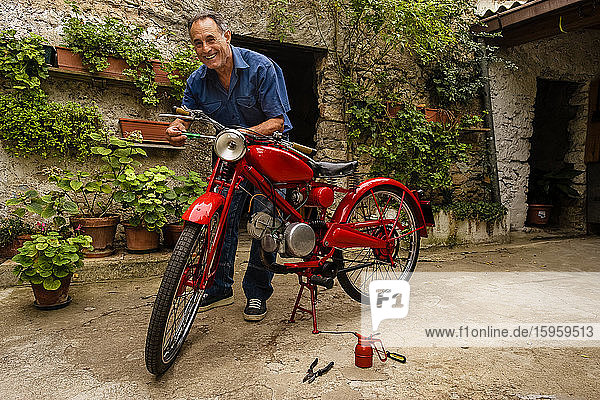 Ein reifer Mann steht im Hof und repariert ein rotes Oldtimer-Motorrad.