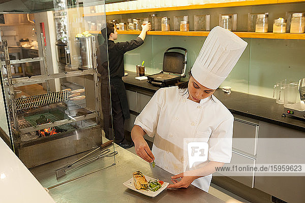 Female chef wearing chef's hat standing at worktop in commercial kitchen  garnishing plate of food.