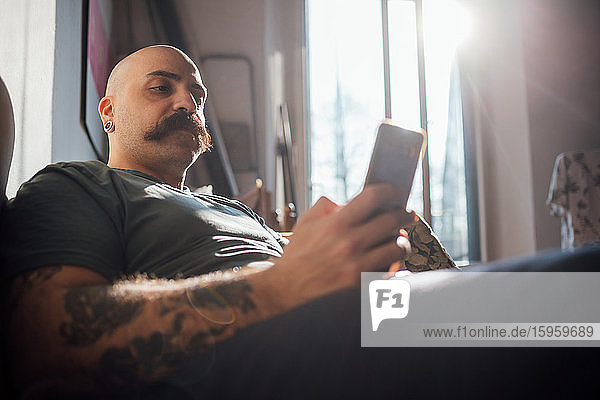Kahlköpfiger Mann mit Schnurrbart auf dem Sofa liegend  während der Corona-Krise mit dem Handy telefonierend und sich selbst isolierend.