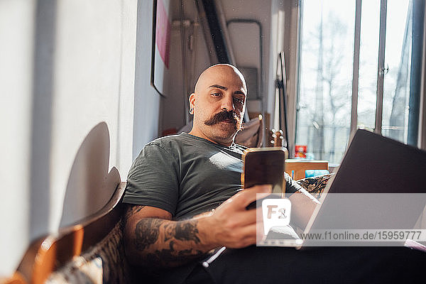 Kahlköpfiger Mann mit Schnurrbart auf dem Sofa liegend  während der Corona-Krise mit dem Handy telefonierend und sich selbst isolierend.