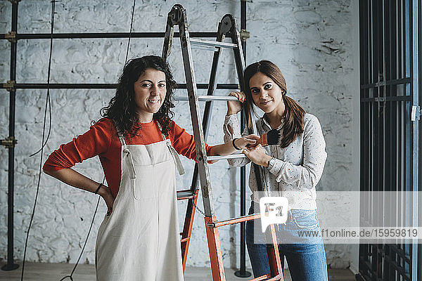 Zwei mittelgroße erwachsene Frauen  die sich in ihrem neuen Laden auf Trittleitern mit Pinsel abstützen  Porträt