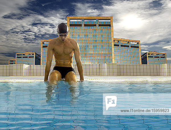 Man wearing black swimming trunks  cap and goggles sitting on edge of outdoor swimming pool  modern skyscrapers in background.