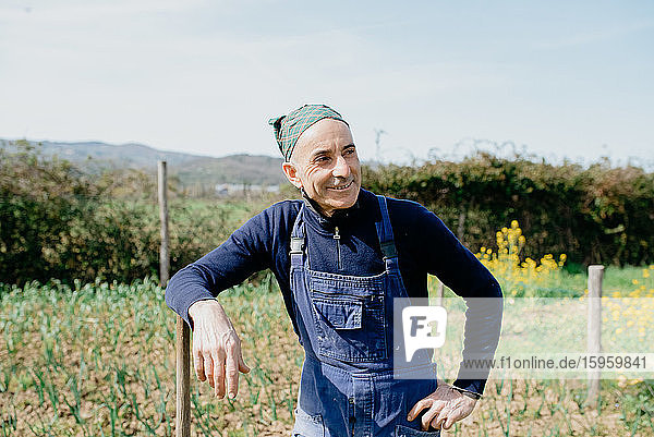 Lächelnder Mann in Latzhose und Kopftuch  im Gemüsegarten stehend  an einer Holzstange gelehnt.