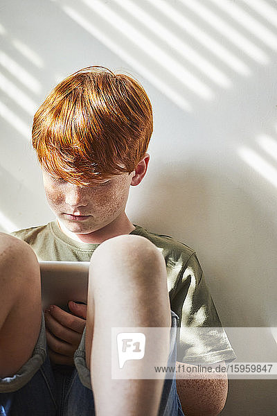 Junge mit roten Haaren sitzt auf dem Boden in einem sonnigen Raum und hält ein digitales Tablett in der Hand.