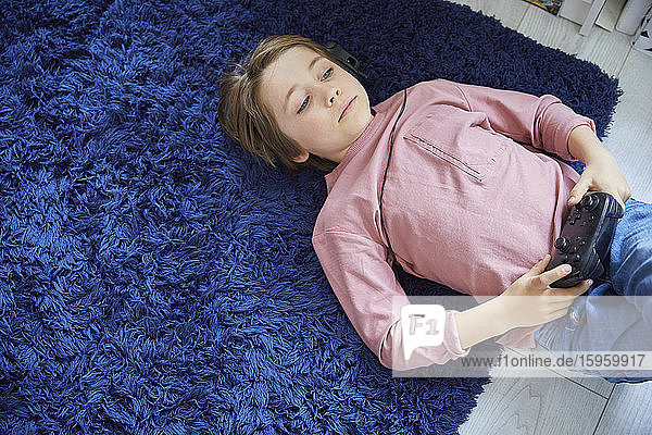 Junge mit rosa T-Shirt auf blauem flauschigen Teppich auf dem Rücken liegend den Controller der Spielkonsole haltend.