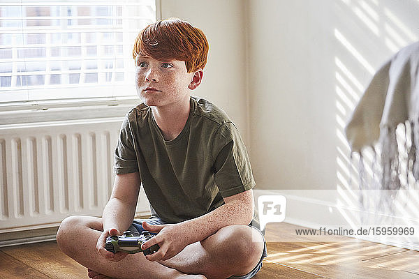 Boy with red hair sitting on floor in sunny room  holding game console controller.