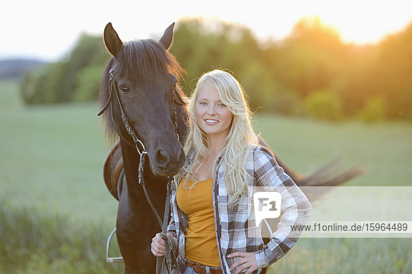 Young woman standing beside a horse on a meadow at sunset