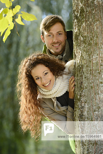 Lächelndes Paar schaut hinter einem Baumstamm hervor