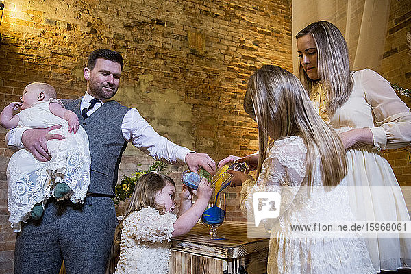 Family pouring coloured sand into glass jar during naming ceremony in an historic barn.