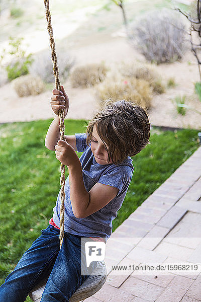 Six year old boy using a rope swing on a wide porch in the shade.
