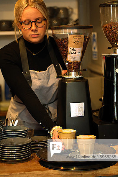 Blonde Frau mit Brille und Schürze steht am Tresen eines Cafés und stellt zwei Café Latte auf ein Tablett.