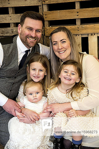 Porträt von lächelnden Eltern mit ihren drei kleinen Töchtern während der Taufzeremonie in einer historischen Scheune.