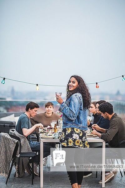 Portrait of smiling woman with drink standing by table during social gathering on terrace building