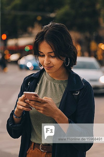 Smiling young female texting through phone while standing in city