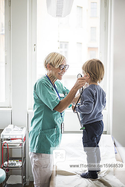 Smiling female doctor holding stethoscope to boy's ear while standing in hospital