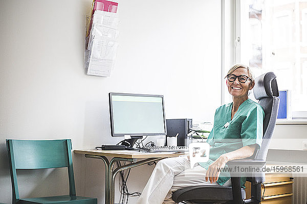 Portrait of smiling mature female doctor sitting on chair at clinic desk