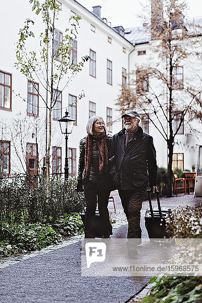 Glückliches älteres Ehepaar geht mit Gepäck auf einem Fußweg vor dem Hotel