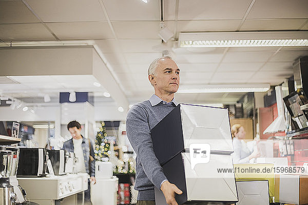 Mature salesman with boxes looking away while walking in electronics store